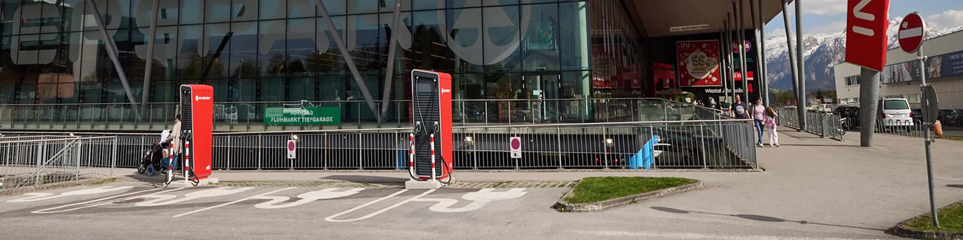 Zwei rote Ladestationen für Elektrofahrzeuge vor einem modernen Glasgebäude mit angebrachten Schildern und schneebedeckten Bergen im Hintergrund.