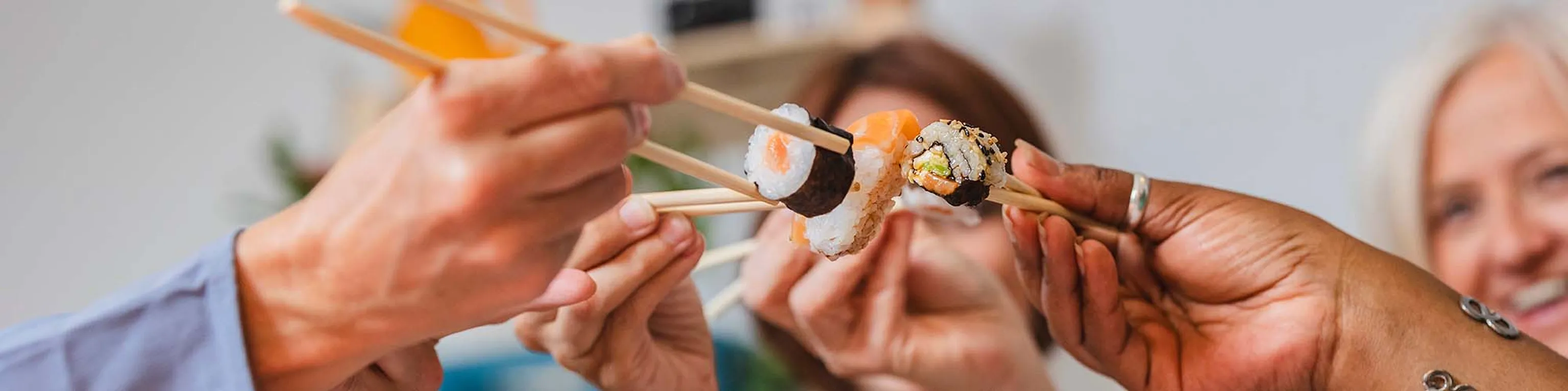 Hände, die Sushi mit Essstäbchen halten und diese feierlich aneinanderklirren lassen, im Hintergrund verschwommene Gesichter.