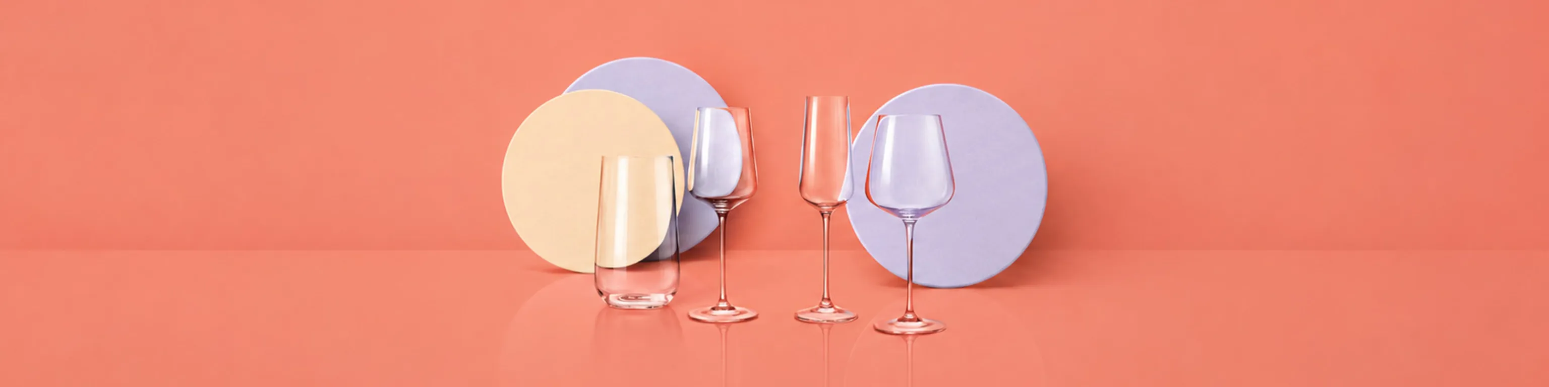 Mehrere verschiedene Glas-Produkte wie Weingläser, Wassergläser und Teller von Spiegelau vor einem lachsfarbenen Hintergund