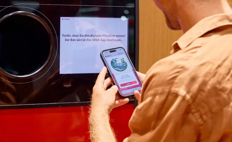 Eine Person, die vor einem Pfandautomaten mit einer Smartphone-App über einen digitalen Bildschirm wischt. Auf dem Bildschirm ist ein deutscher Text zu sehen.
