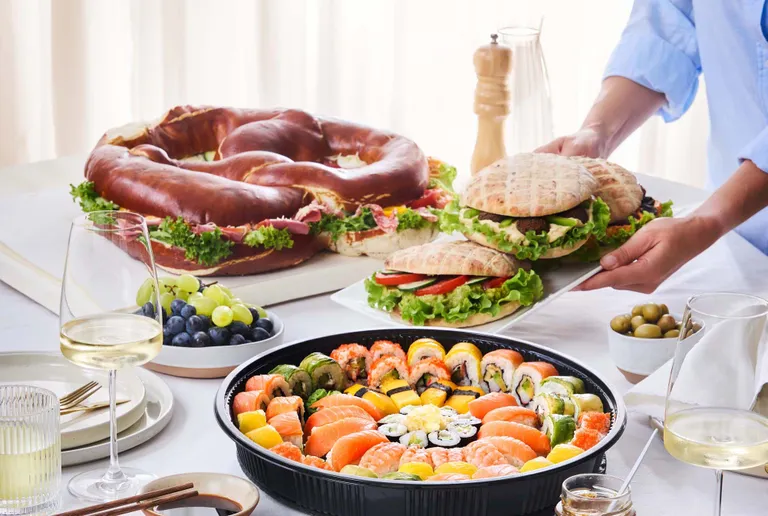 Ein Tisch, gedeckt mit einem riesigen Brezel-Sandwich, einer bunten Sushi-Platte, Pitas, Weintrauben, Oliven und Gläsern mit Weißwein.