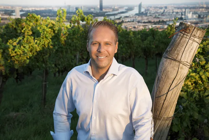 Ein Mann in einem weißen Hemd steht lächelnd in einem Weinberg, im Hintergrund sind in der Ferne eine Stadtlandschaft und ein Fluss zu sehen.