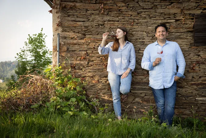 Ein Mann und eine Frau lehnen an einer rustikalen Steinmauer, jeder hält ein Glas Rotwein in der Hand, sie sind von Grün umgeben und tragen legere blaue Hemden.