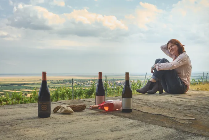 Eine Frau sitzt auf einer Terrasse, neben ihr stehen drei Weinflaschen; im Hintergrund erstreckt sich unter bewölktem Himmel eine malerische Weinberglandschaft.