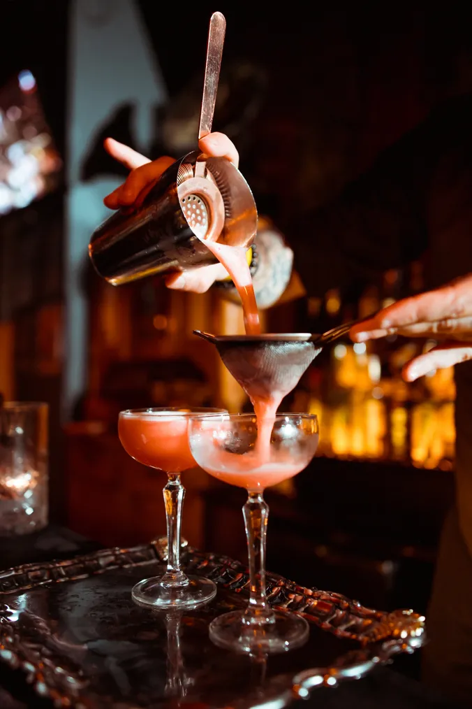 Ein Barkeeper gießt einen rosa Cocktail durch ein Sieb in Coupé-Gläser, vor einem scharfgestellten Hintergrund und bei warmer Beleuchtung.