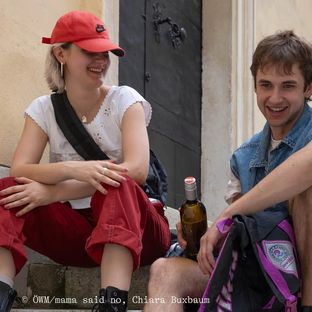 Two people sitting on steps, smiling and talking. One wears a red cap and white top; the other in a denim vest holds a bottle.