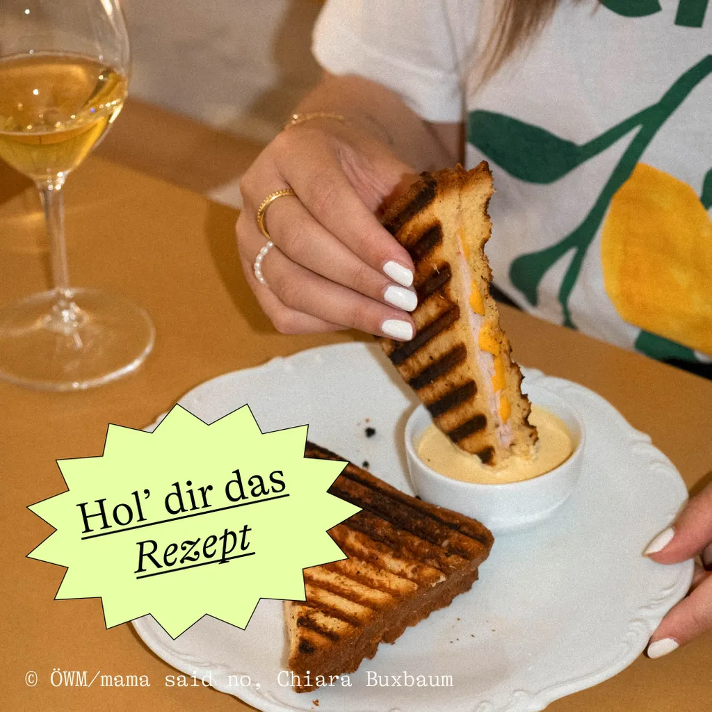 A person dips a grilled sandwich into sauce on a plate, with a wine glass nearby and text overlay "Hol’ dir das Rezept."