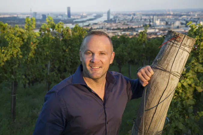 Ein Mann in einem blauen Hemd steht lächelnd in einem Weinberg, im Hintergrund ist die Skyline einer Stadt zu sehen.