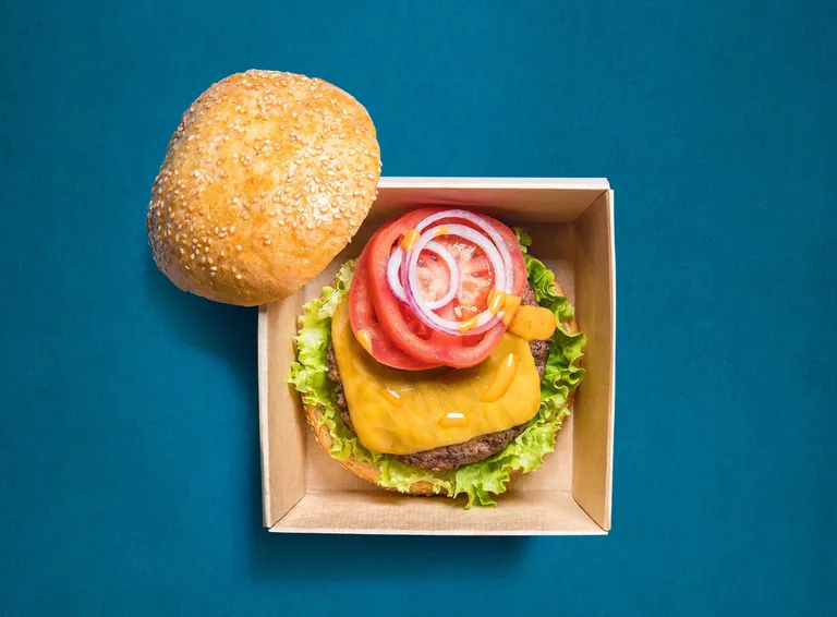 Ein offener Burger in einer Schachtel vor blauem Hintergrund, mit Salat, Käse, Tomate und Zwiebel, daneben die obere Hälfte eines Sesambrötchens.
