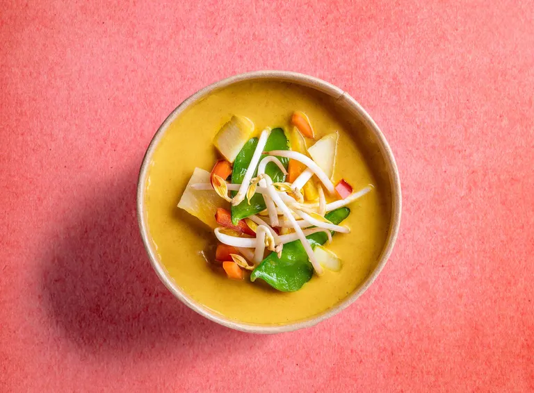 Eine Schüssel gelbes Curry mit buntem Gemüse und Sojasprossen vor einem rosa Hintergrund.