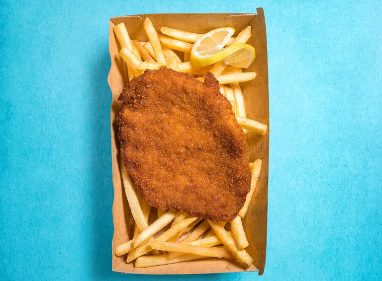 Ein paniertes Schnitzel auf knusprigen Pommes frites in einer Papierschale, garniert mit Zitronenscheiben, vor einem leuchtend blauen Hintergrund.