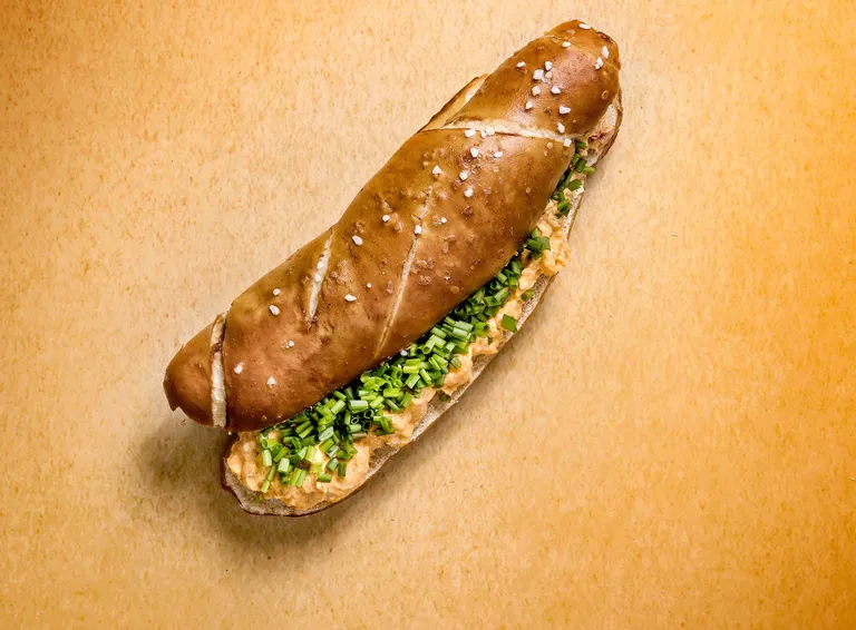 Ein Laugenstangerl mit cremiger Füllung und gehacktem Schnittlauch, bestreut mit grobem Salz, auf einer hellbraunen Unterlage.
