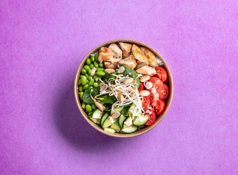 Eine farbenfrohe Bowl mit gegrilltem Hähnchenfleisch, Edamame, Gurken, Kirschtomaten, Sprossen und Mandelscheiben vor einem rosa Hintergrund.