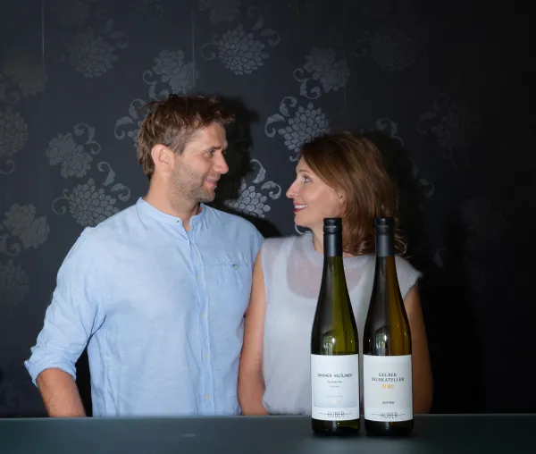 Eine Frau und ein Mann stehen gegenüber und blicken sich ins Gesicht und im Vordergrund stehen zwei Weinflaschen des Weingut Markus Huber auf einem Tisch.