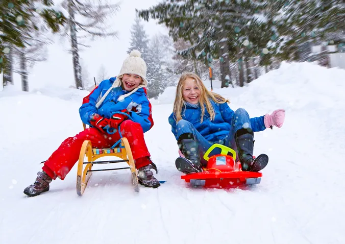 Kinder auf einer Rodel und einem Bob, auf einer Piste im Winter, verschneite Bäume im Hintergrund
