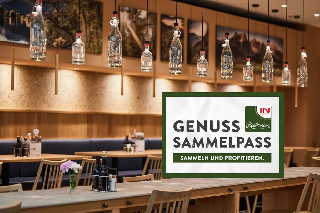 Modernes Restaurantinterieur mit Holztischen und hängenden Flaschen. Im Vordergrund ist ein Schild mit der Aufschrift „GENUSS SAMMELPASS – SAMMELN UND PROFITIEREN“ zu sehen.