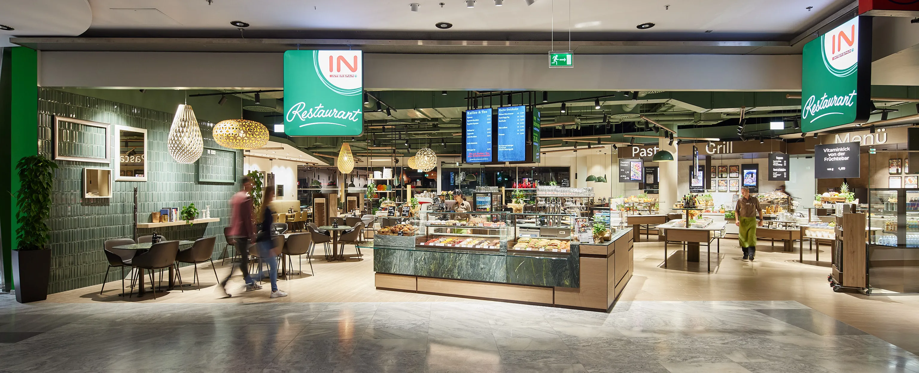Modernes Restaurant mit Sitzgelegenheiten, einer Auslage für Backwaren und Menübildschirmen an der Decke. Heller Raum mit Hängeleuchten und grünen Akzenten.