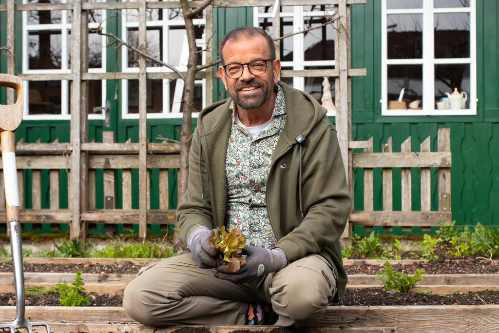 Ein lächelnder Mann in einem Garten, der auf einem hölzernen Hochbeet sitzt, einen Salatkopf in der Hand hält, eine Brille und Gartenhandschuhe trägt, im Hintergrund sind grüne Türen zu sehen.