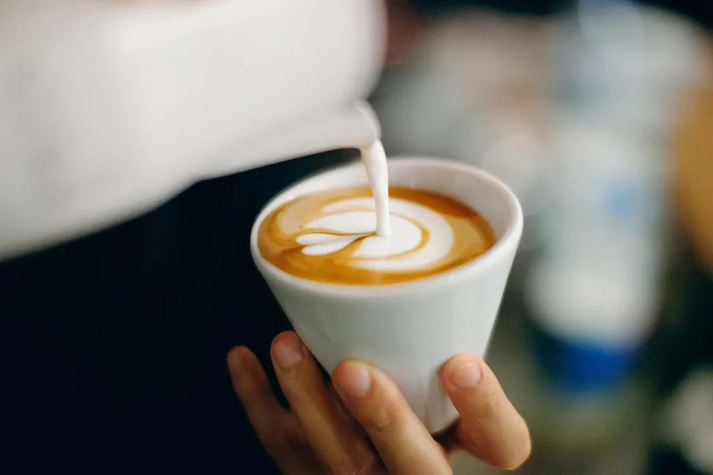 Nahaufnahme einer Hand, die eine Tasse Cappuccino hält, während Milch eingegossen wird und sich auf der Oberfläche Latte-Art bildet.