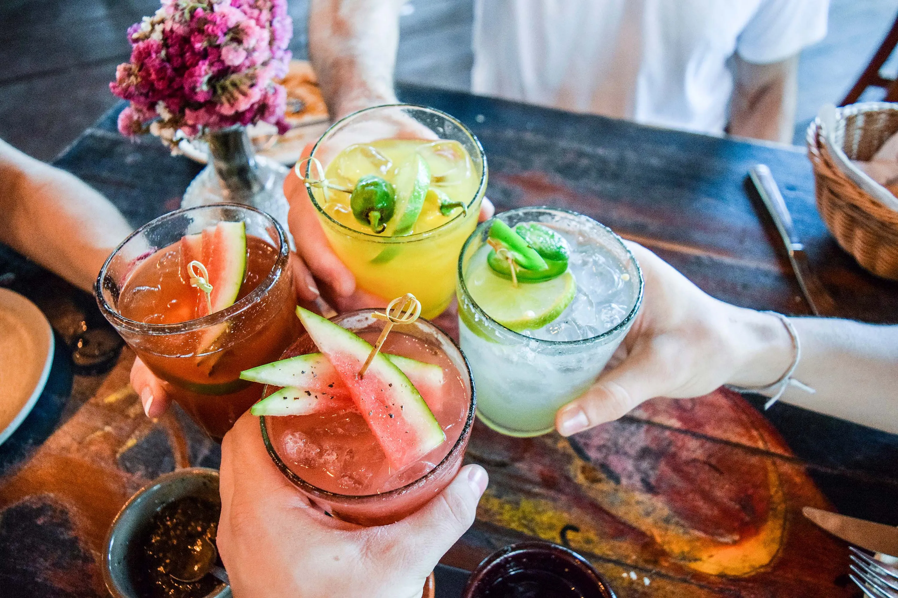Vier farbenfrohe Cocktails, garniert mit Früchten und Jalapeños, werden an einem Holztisch zum Anstoßen aneinandergestoßen.