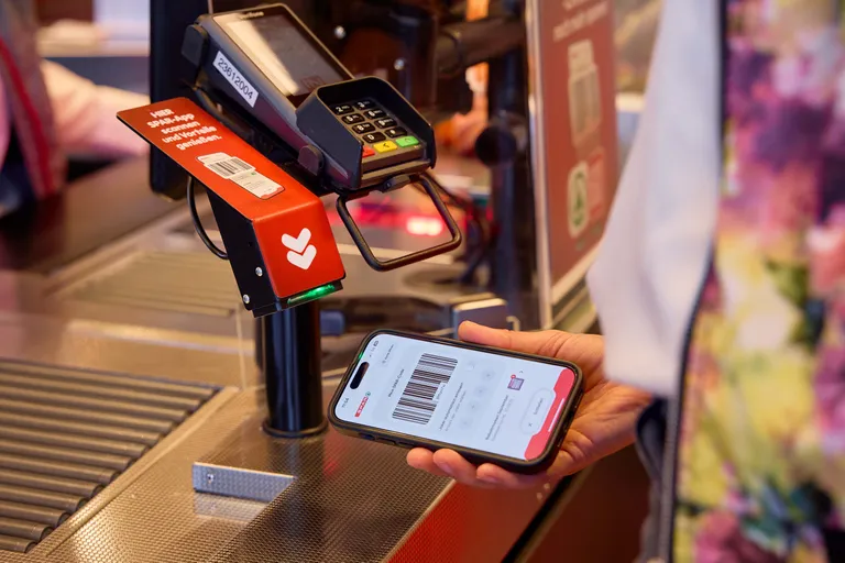 Eine Person scannt mit ihrem Smartphone einen Barcode unter dem Scanner an der Ladentheke. Auf dem Handy sind ein digitaler Code und eine Zahlungs-App zu sehen.