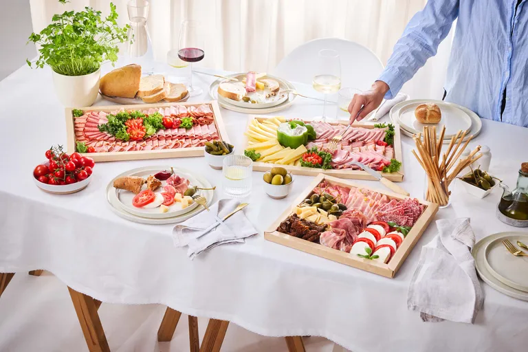 Eine elegante Tischdekoration mit verschiedenen Wurst- und Käsesorten, Brot, Oliven, Tomaten und Wein. Eine Hand greift nach einer Scheibe Wurst.