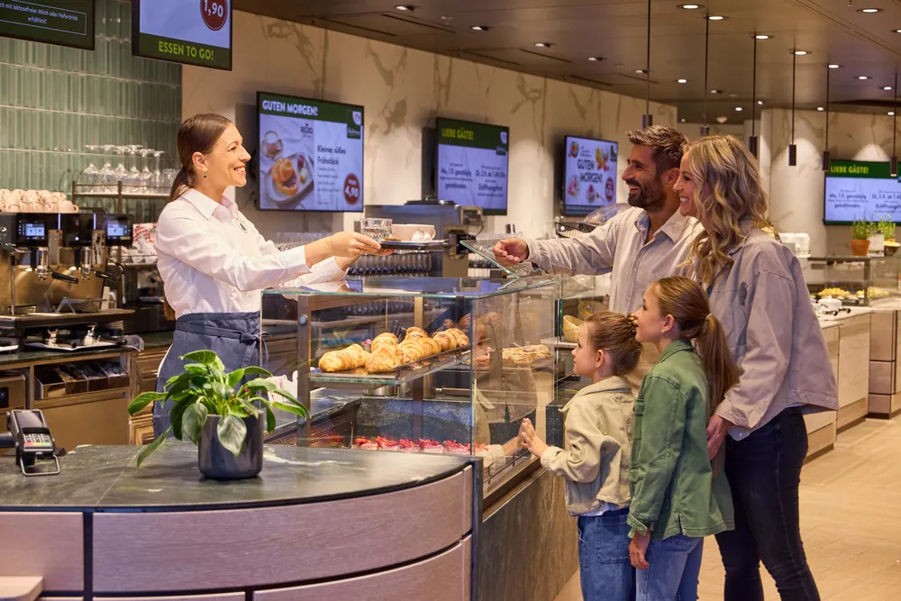 Eine Familie bestellt Gebäck bei einer freundlich lächelnden Verkäuferin an der Theke einer Bäckerei, in deren Vitrine eine Vielzahl von Backwaren ausgestellt ist.