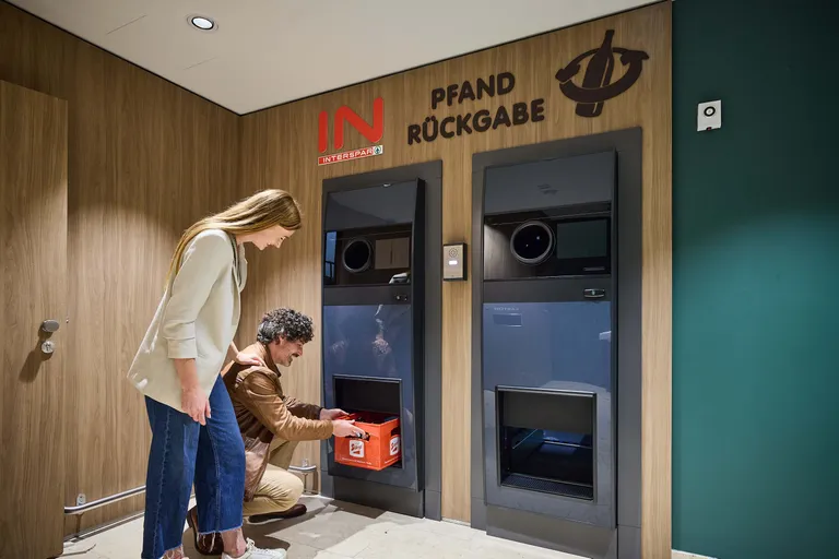 Ein Mann und eine Frau nutzen einen Flaschenrücknahmeautomaten mit der Aufschrift „Pfand Rückgabe“ in einem holzgetäfelten Raum.