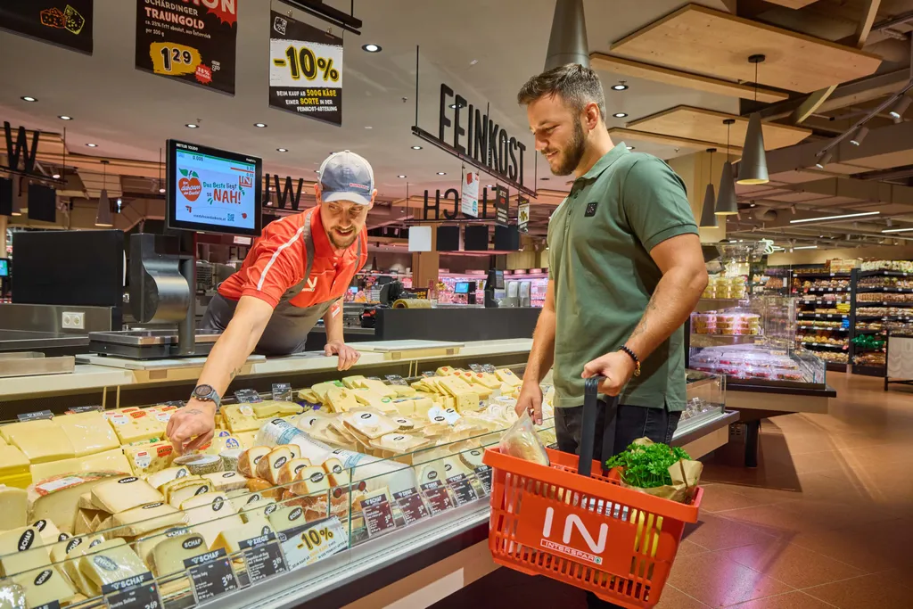 Ein Kunde und ein Mitarbeiter an der Käsetheke eines Supermarkts. Der eine zeigt auf verschiedene Käsesorten, während der andere einen Einkaufskorb mit Lebensmitteln in der Hand hält.