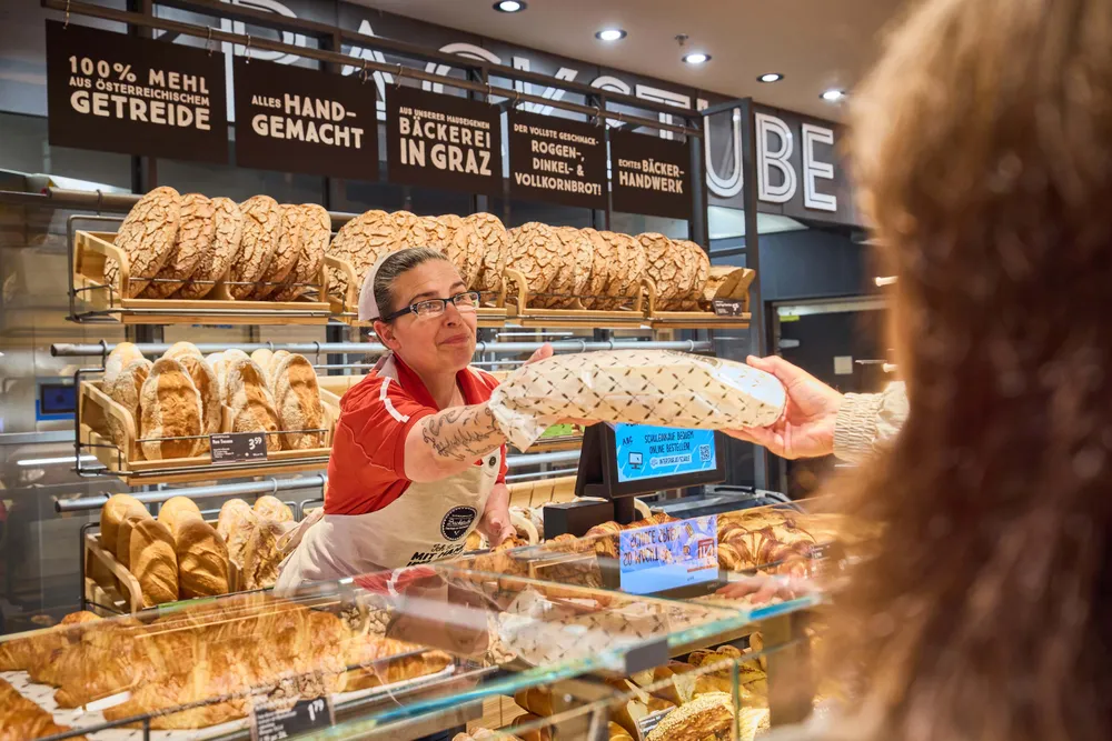 Ein Bäckereimitarbeiter mit Brille reicht einem Kunden einen Laib Brot. Im Hintergrund sind Regale mit verschiedenen Brotsorten zu sehen.