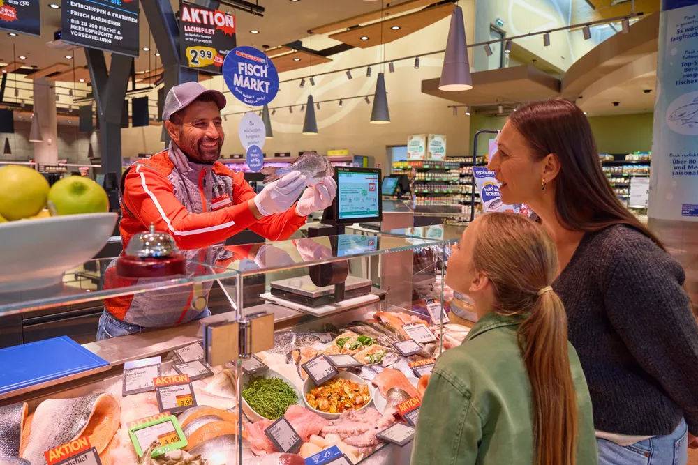 Ein lächelnder Fischhändler mit Mütze zeigt einer Frau und einem Kind an der Fischtheke eines Supermarkts einen Fisch.