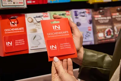 Eine Person hält in einem Geschäft inmitten verschiedener Karten eine rote Geschenkkarte der Marke „IN“ in der Hand, auf der „Das perfekte Geschenk für jeden Anlass!“ steht.