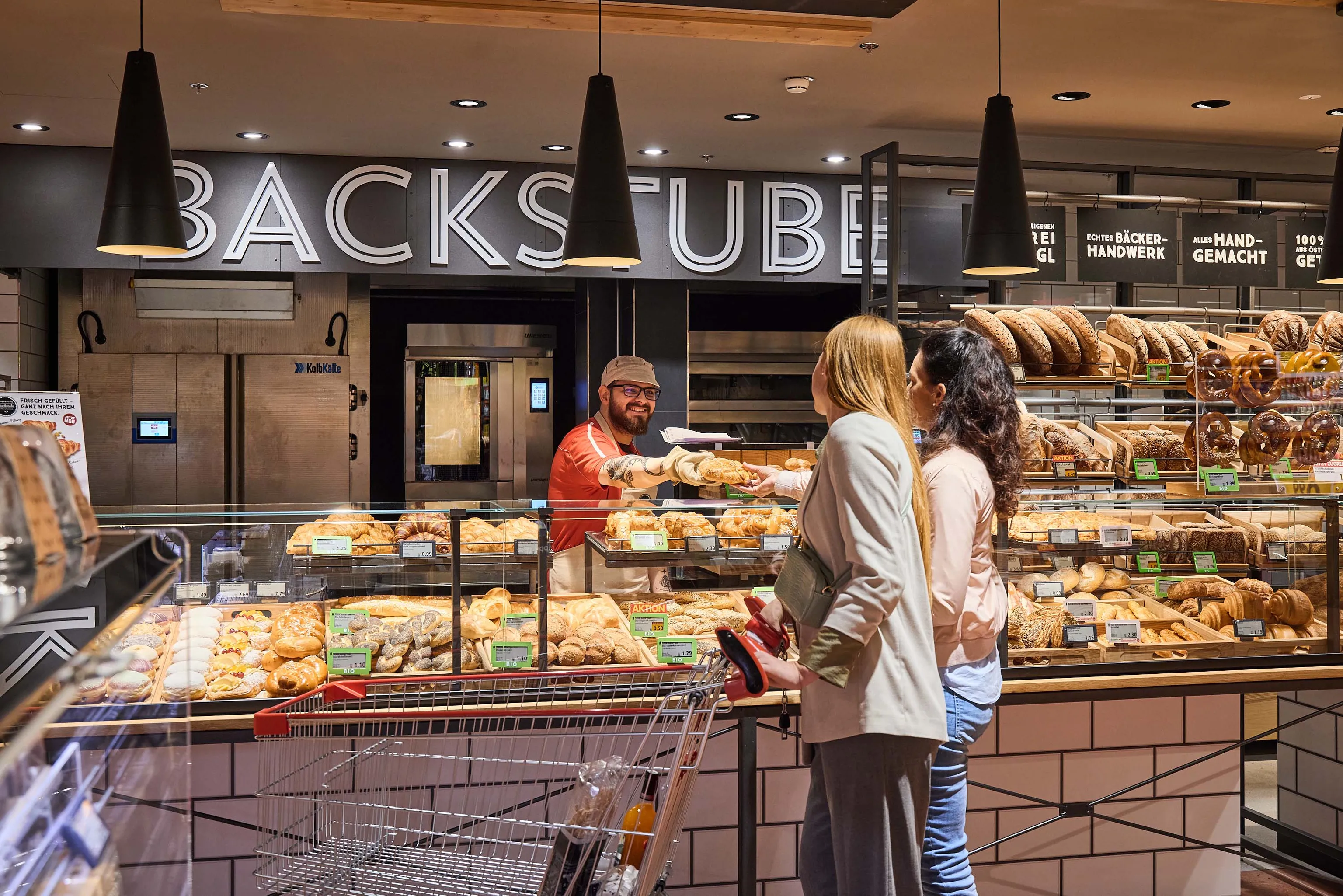 Zwei Frauen kaufen Gebäck bei einer freundlich lächelnden Verkäuferin an der Bäckereitheke in einem Supermarkt, umgeben von verschiedenen Brotsorten und Backwaren.