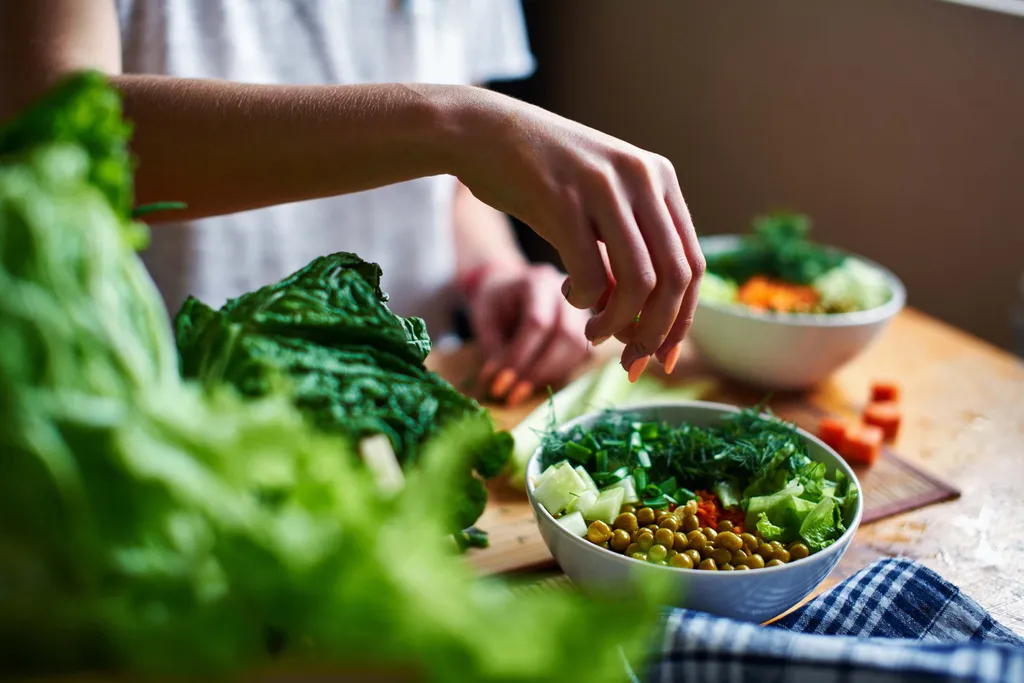 Eine Person bereitet in einer Schüssel auf einem Holztisch einen frischen Salat aus Blattgemüse, Erbsen, Gurken und Kräutern zu.