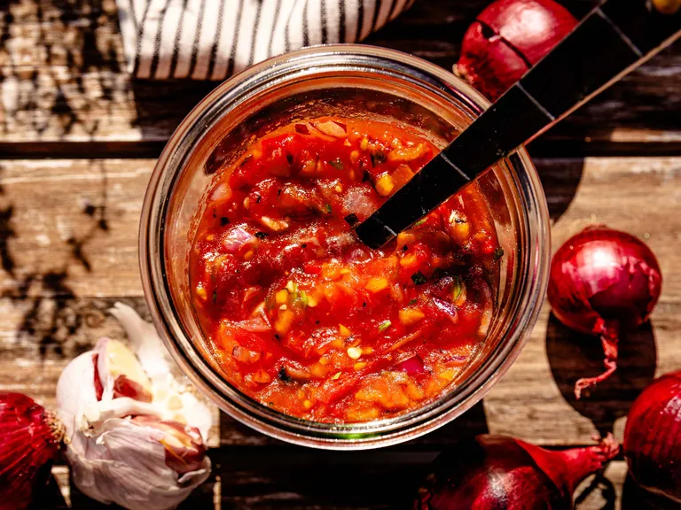 Eine rote Salsa mit einem Löffel in einem Einweckglas das auf einem Holzbrett auf dem Zwiebeln liegen