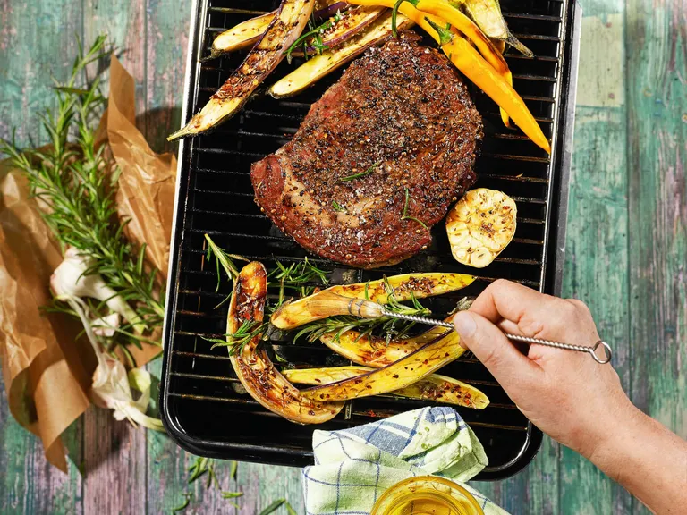 Eine Person pinselt gerade Zucchini-Scheiben mit Barbecue-Sauce auf einem Grill auf dem auch ein Rumpsteak liegt ein