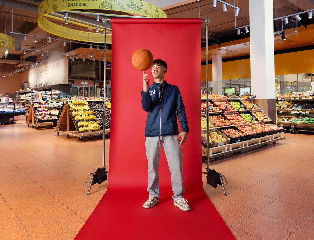 Lehrling lässt einen Basketball auf dem Finger kreisen und steht vor rotem Fotohintergrund im Markt