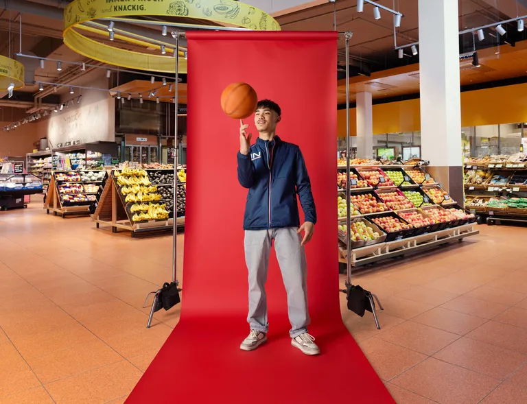 Lehrling lässt einen Basketball auf dem Finger kreisen und steht vor rotem Fotohintergrund im Markt