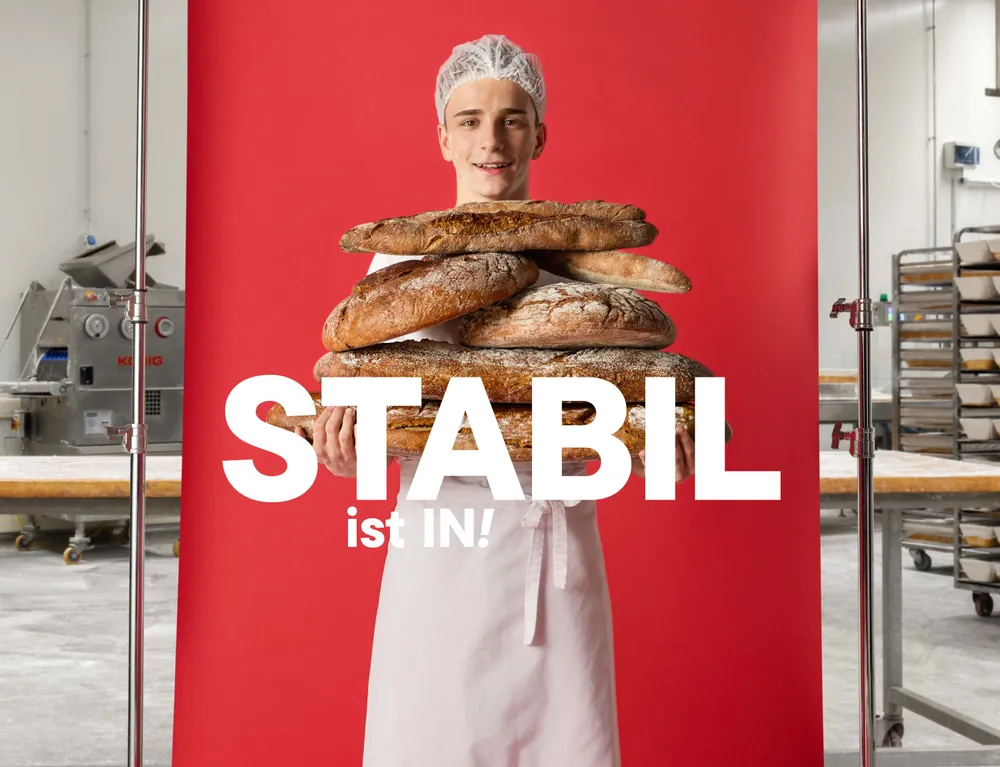 Junger Mitarbeiter in einer Backstube trägt mehrere Brote vor rotem Hintergrund mit dem Schriftzug „Stabil ist IN!“