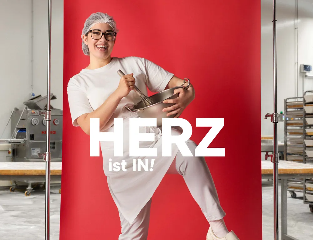 Junge Mitarbeiterin in einer Backstube mit Rührschüssel vor rotem Hintergrund und dem Schriftzug „Herz ist IN!“