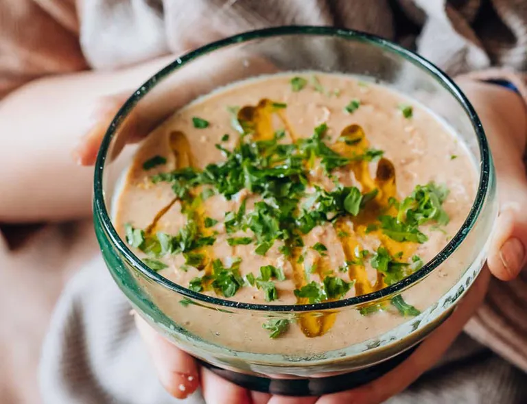 Frau hält eine Schale mit cremiger Suppe, frischen Kräutern und Öl in den Händen