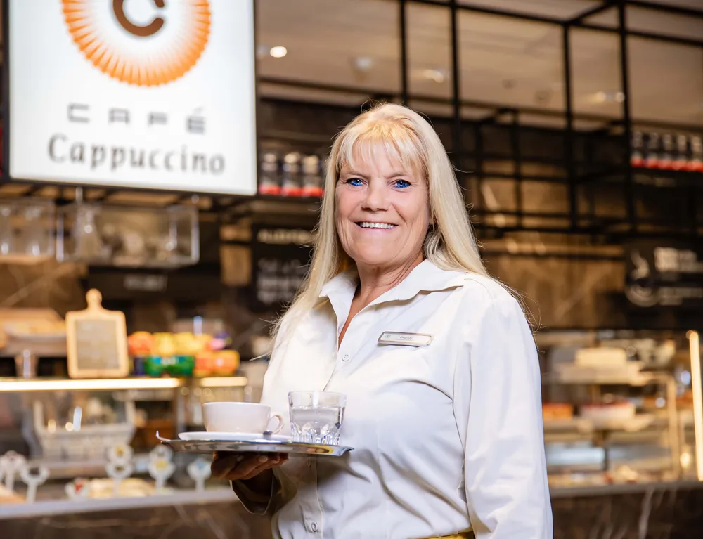 Lächelnde Mitarbeiterin im INTERSPAR Café Cappuccino mit Kaffee und Wasser auf einem Tablett