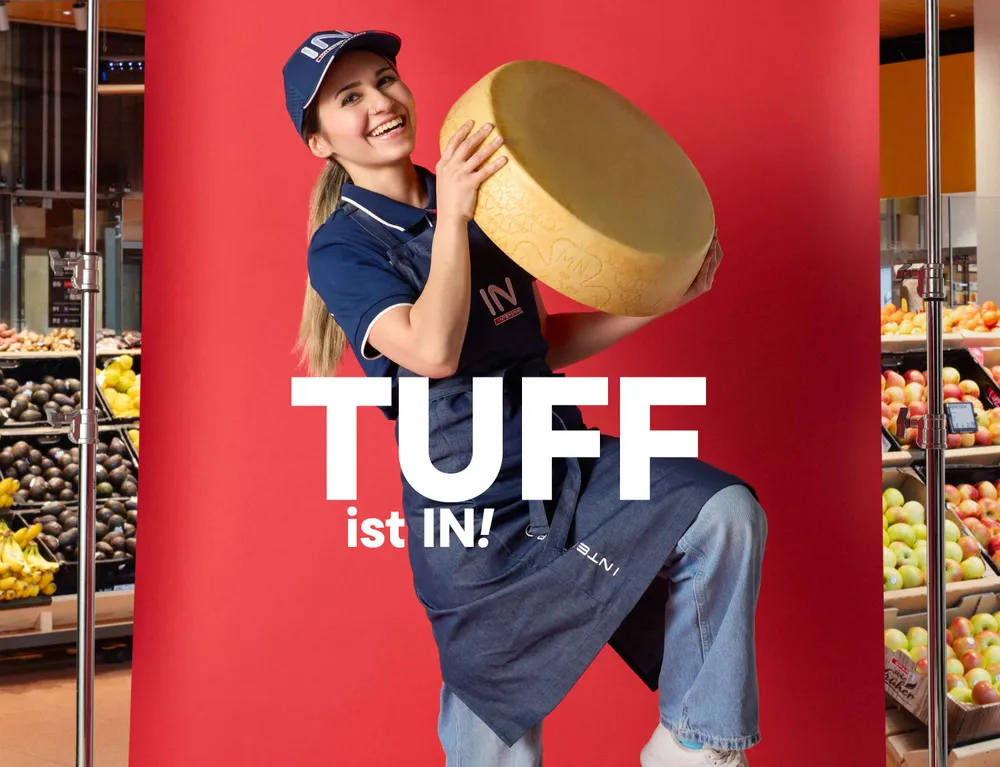 Junge INTERSPAR Mitarbeiterin hält einen großen Käselaib vor rotem Hintergrund mit dem Schriftzug „Tuff ist IN!“