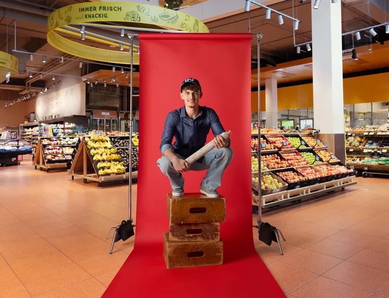 Lehrling im Feinkostbereich sitzt mit einem Produkt auf Holzkisten vor rotem Fotohintergrund im INTERSPAR-Markt