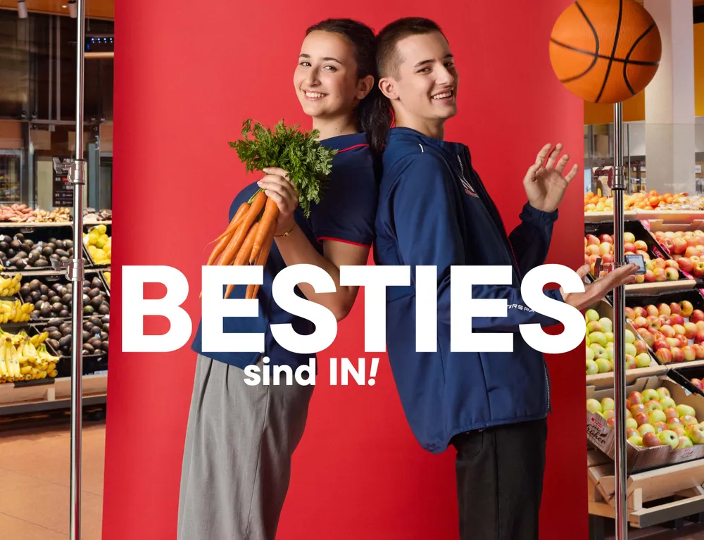 Zwei junge INTERSPAR Mitarbeitende mit Karotten und Basketball vor rotem Hintergrund mit dem Schriftzug „Besties sind IN!“
