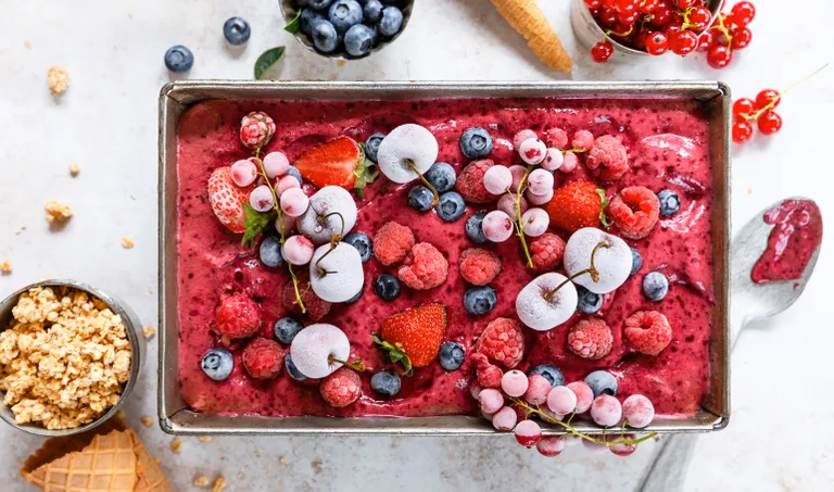Bunter Beeren-Frozen-Yogurt, garniert mit Erdbeeren, Heidelbeeren, Himbeeren und glasierten Kirschen, umgeben von Müsli und einer Waffeltüte.