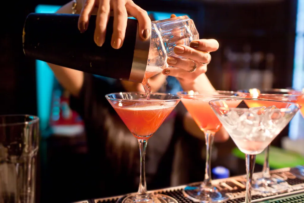 Ein Barkeeper schenkt einen rosa Cocktail in ein Martini-Glas, das neben anderen mit Getränken und Eis gefüllten Gläsern auf einer Bartheke steht.