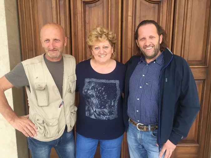 Drei Menschen, die vor einer Holztür lächeln: ein Mann in einer beigen Weste, eine Frau in einem blauen Hemd und ein Mann in einer blauen Jacke.