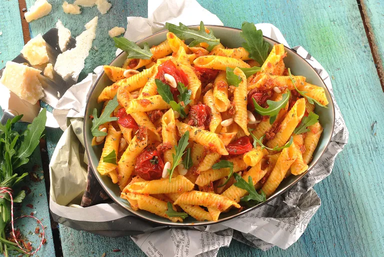 Eine Schüssel Pasta mit Kirschtomaten, Rucola und Pinienkernen auf einem Holztisch, umgeben von geriebenem Käse.
