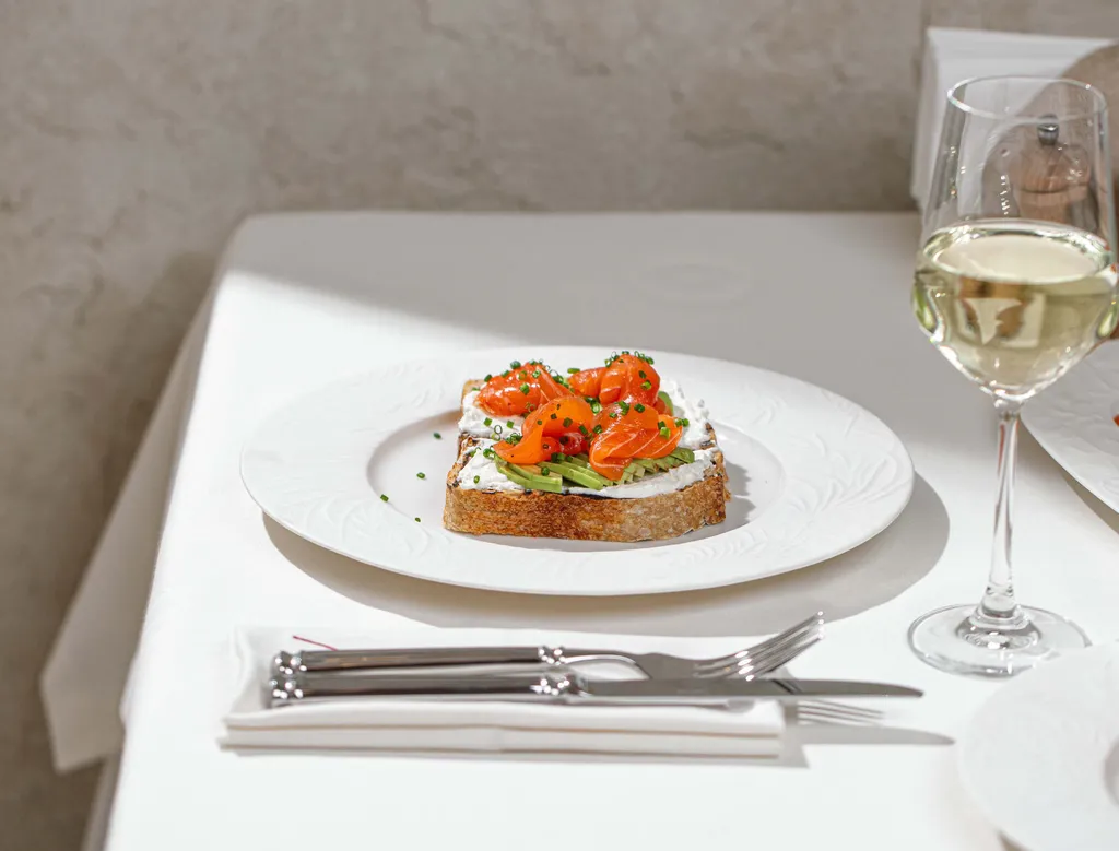 Ein edles Tischgedeck mit einem Glas Weißwein und einem weißen Teller auf dem sich ein Lachs-Sandwich mit Frischkäsecreme befindet